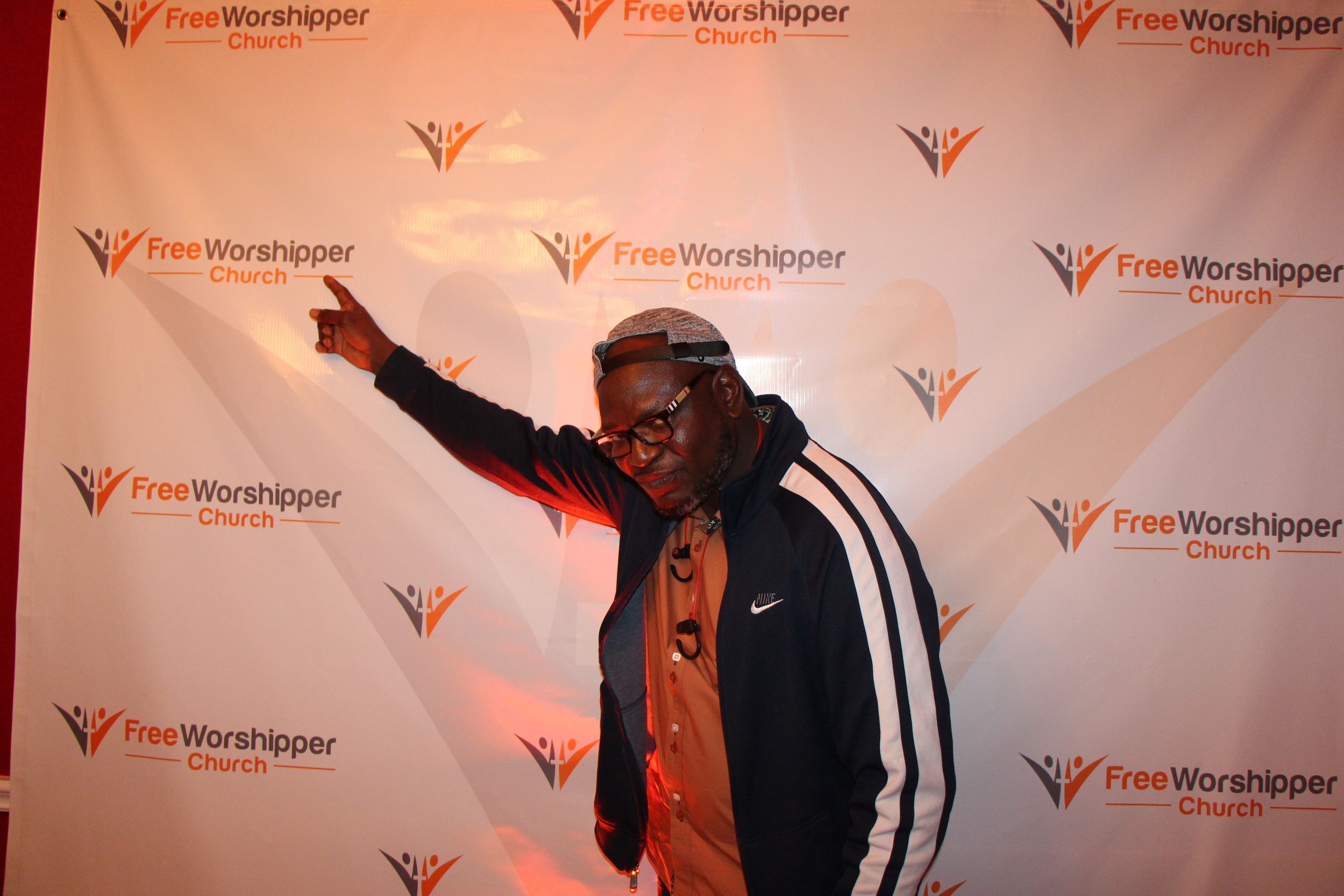 Man posing and pointing in front of the Free Worshipper Church backdrop